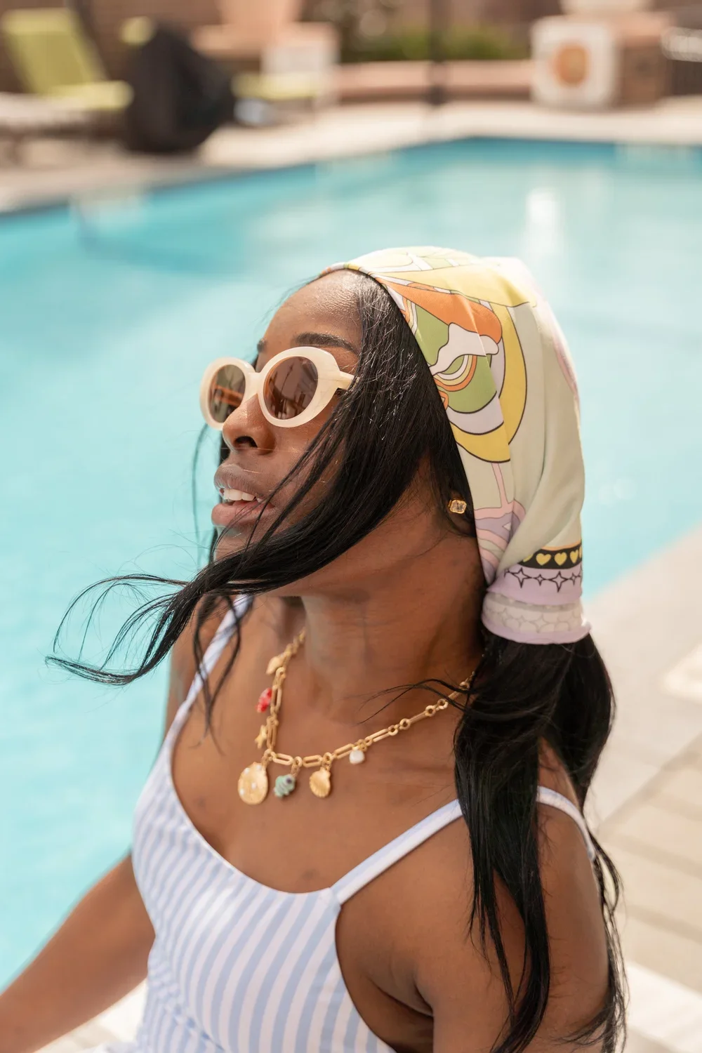 A woman with dark hair wearing sunglasses, a patterned headscarf, and gold jewelry by a swimming pool.
