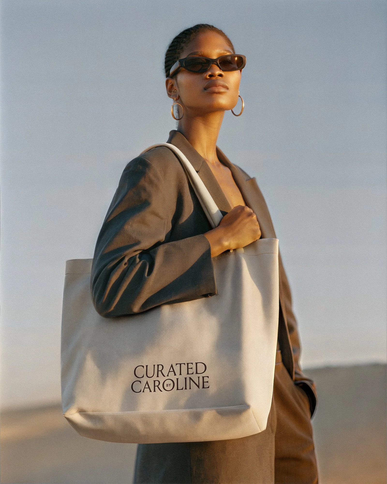 A woman wearing sunglasses and hoop earrings carrying a beige tote bag with the text 'Curated Caroline' on it, outdoors during sunset.