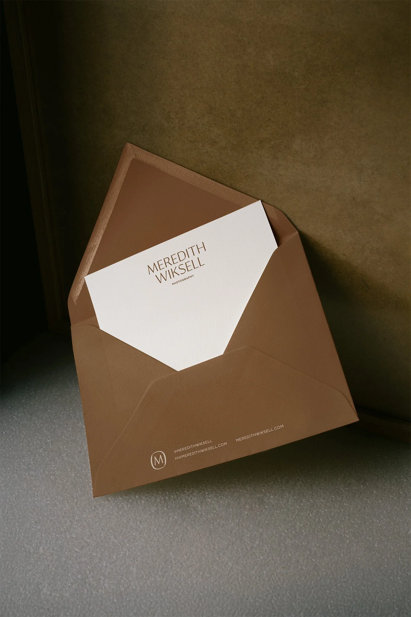 A brown envelope with a white card inside, displaying the name 'Meredith Wikell Photography' in brown text, and contact information at the bottom.
