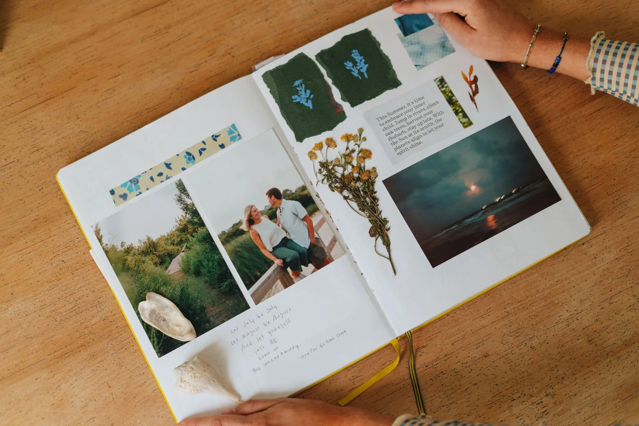 Open scrapbook or photo album showing two photographs of a couple outdoors, dried flowers, a seashell, a handwritten note, and various cutouts and images including a moonlit beach scene, plant illustrations, and a small blue patterned strip at the top.