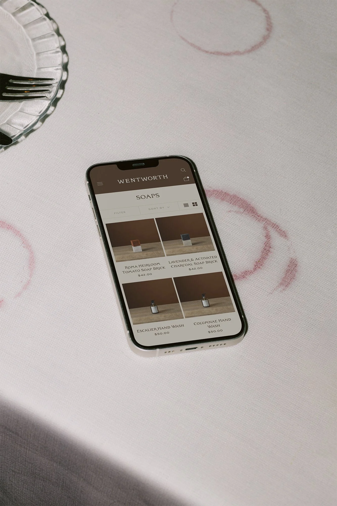 A smartphone on a white table displays an online store for soap products, with various soap options and prices listed. The table also has a glass dish with forks and a pink patterned design.