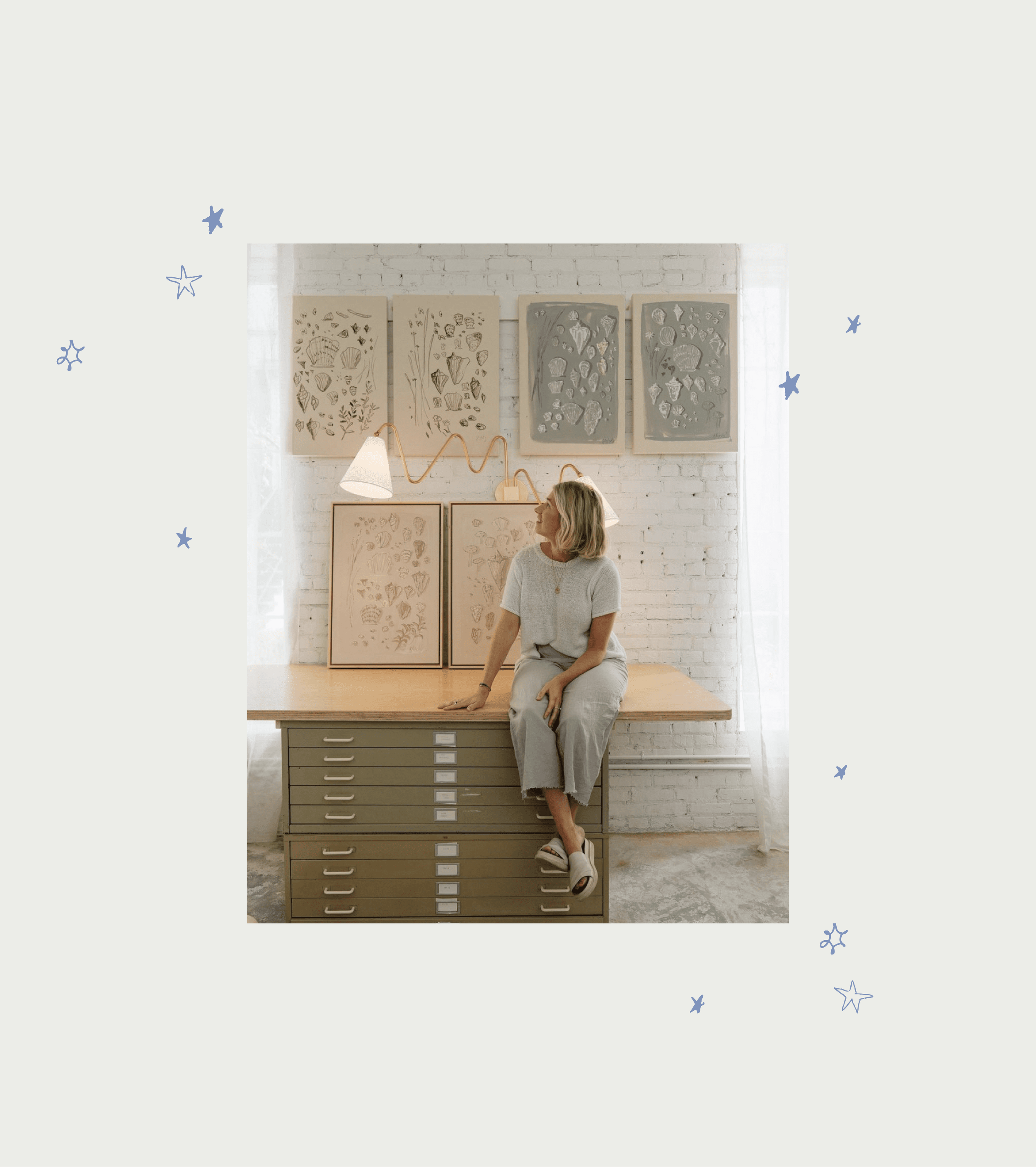 A woman sitting on a filing cabinet in an art studio with artwork behind her and soft natural light.