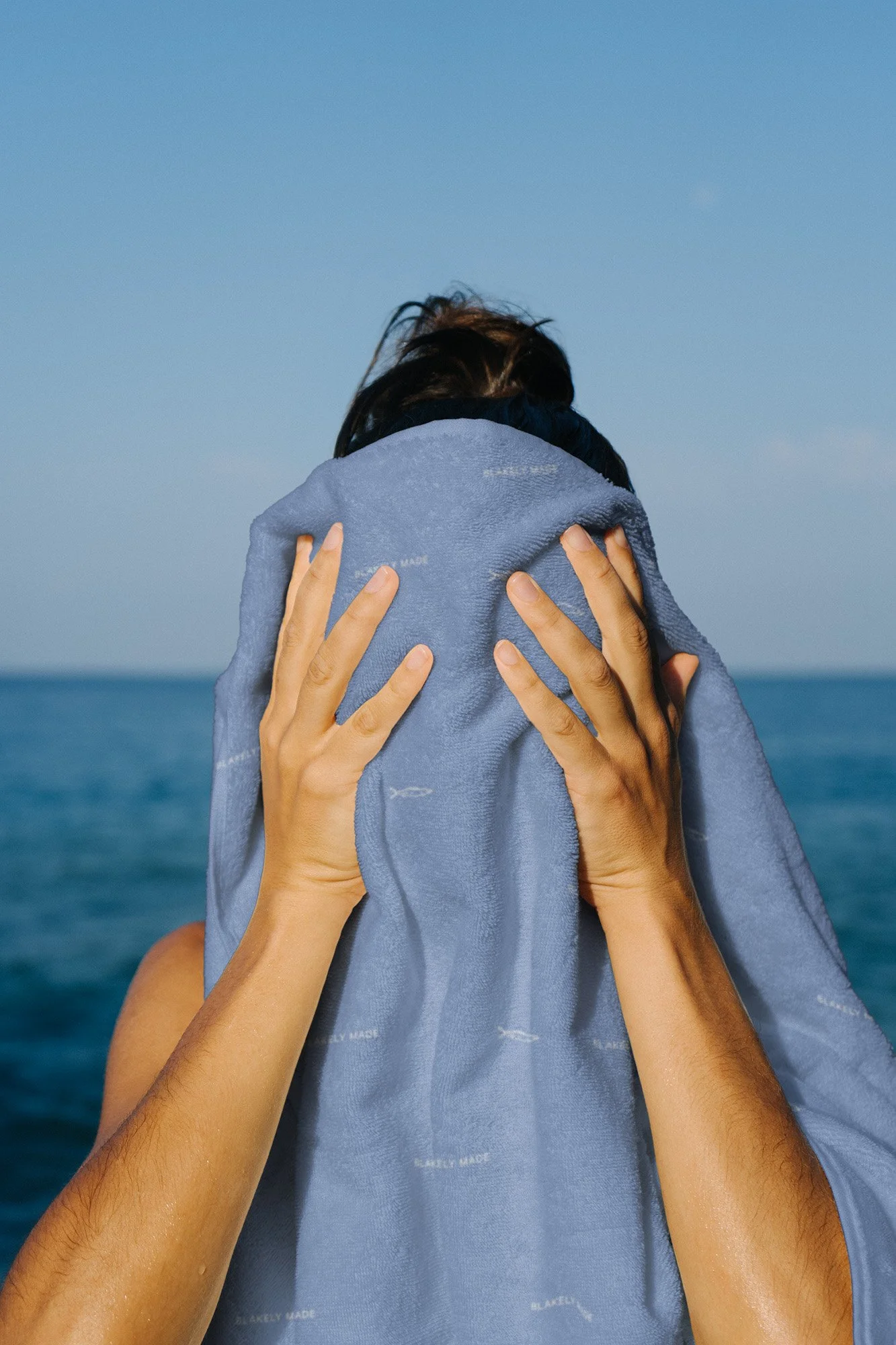 Person covering face with a blue towel against the ocean and clear sky background.