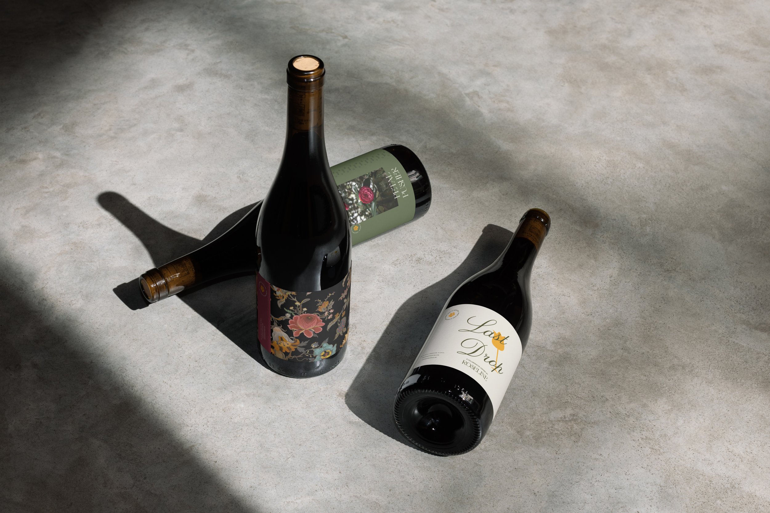 Three wine bottles with floral and text labels on a concrete floor, casting shadows.