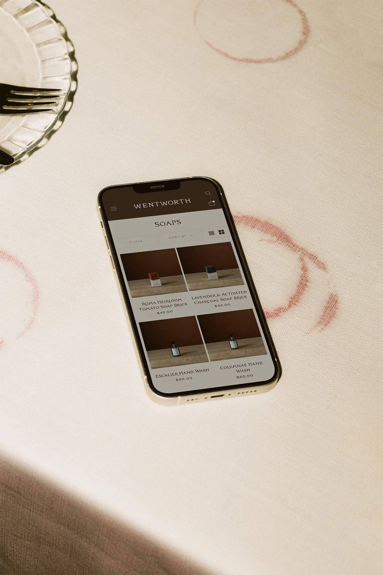 Smartphone displaying an online soap shop on a white table with lipstick marks, a small bowl with a fork and knife, and part of a plate.