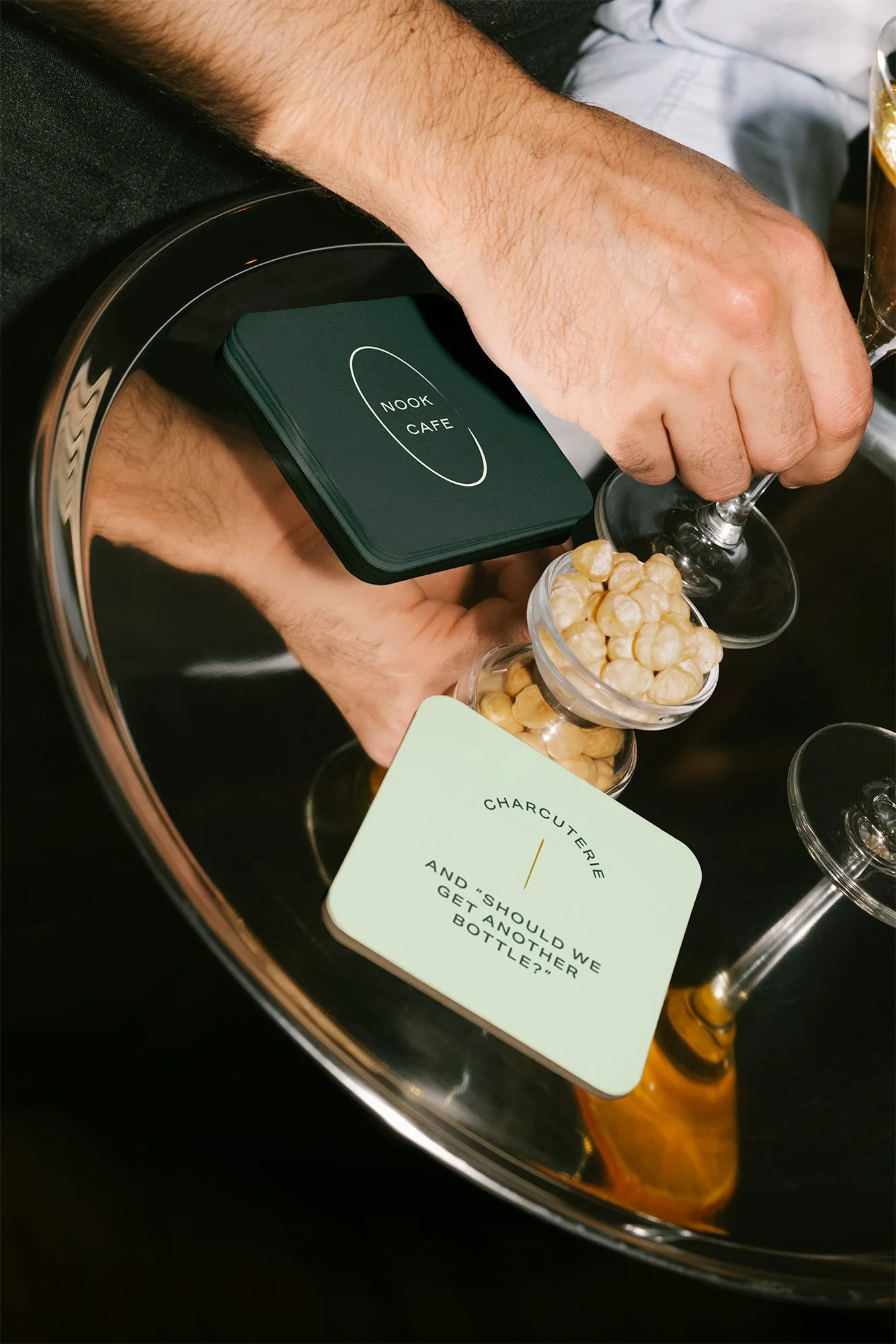 Person's hands placing a card labeled 'Nook Cafe' into a wine bucket filled with chickpeas. A card with words 'Charcuterie and 'Should we get another bottle?' is also in the bucket. A wine glass with a drink is visible in the background.