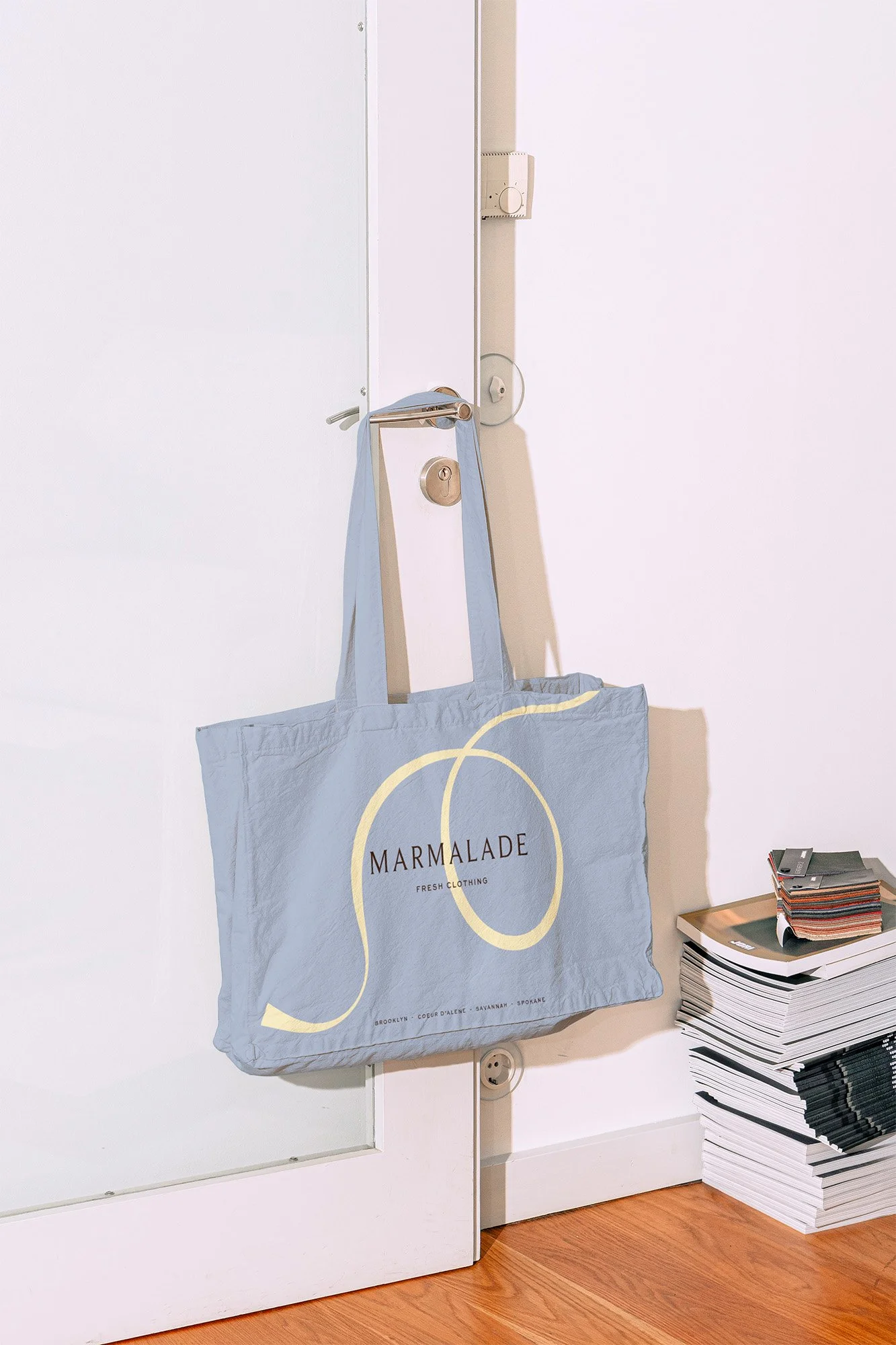 A light blue tote bag labeled 'MARMALADE FRESH CLOTHING' hangs on a door handle. There are stacks of magazines or papers on the floor beside the door.