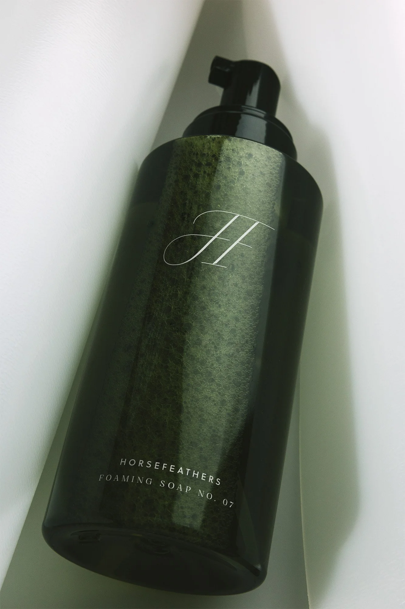 A tall black pump bottle labeled 'Horsefeathers Foaming Soap No. 07' sitting in the corner of a white wall.