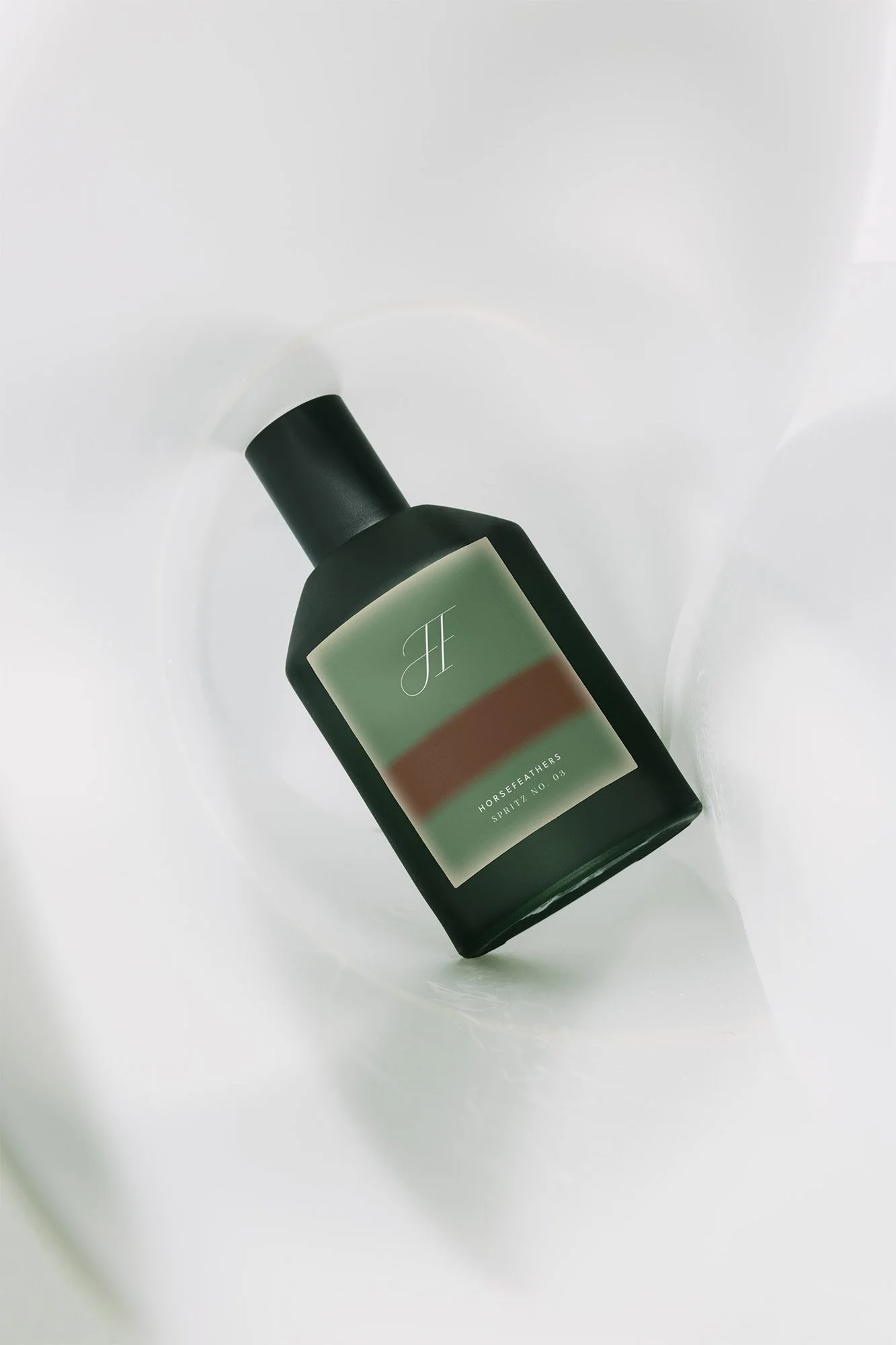 A dark green bottle with a black cap lying on a white surface, featuring a minimalist label with a stylized letter 'H' and text that reads 'Horsefeathers Spirit No. 03'.