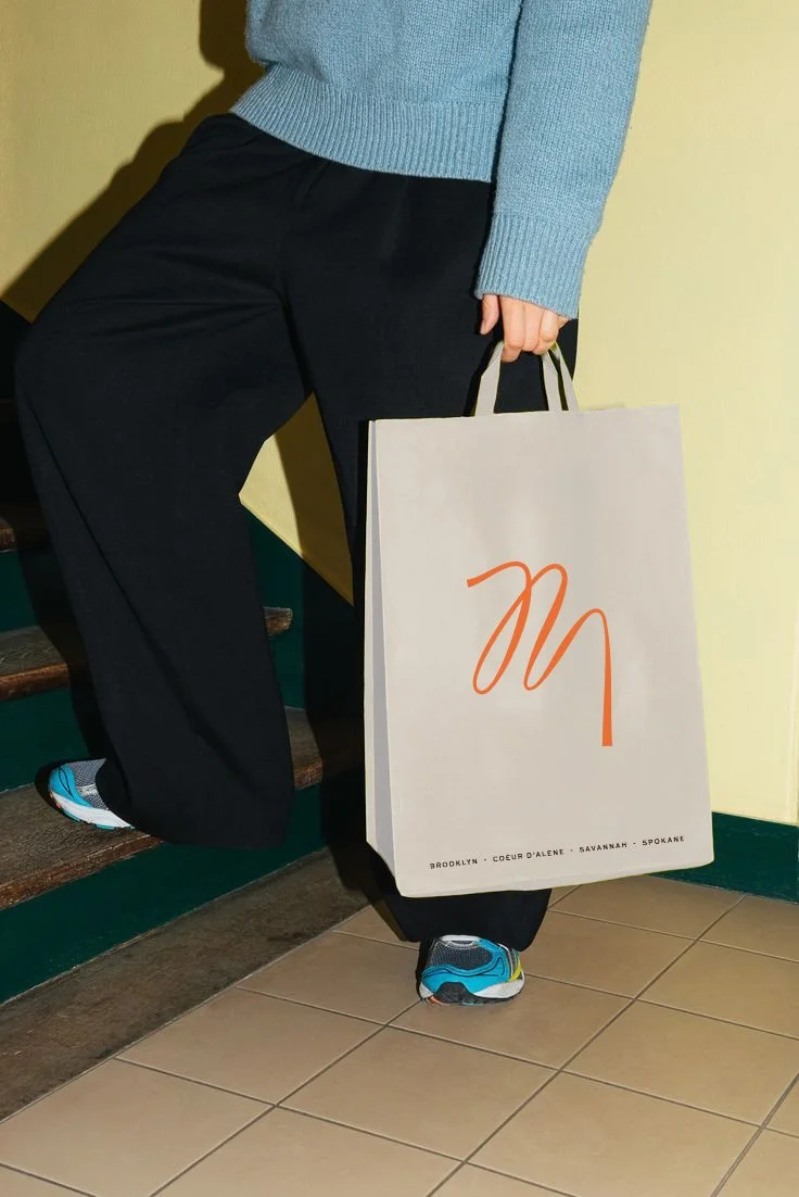 A person in black pants and a blue sweater holding a shopping bag with minimal orange design, standing near stairs in a tiled floor area.