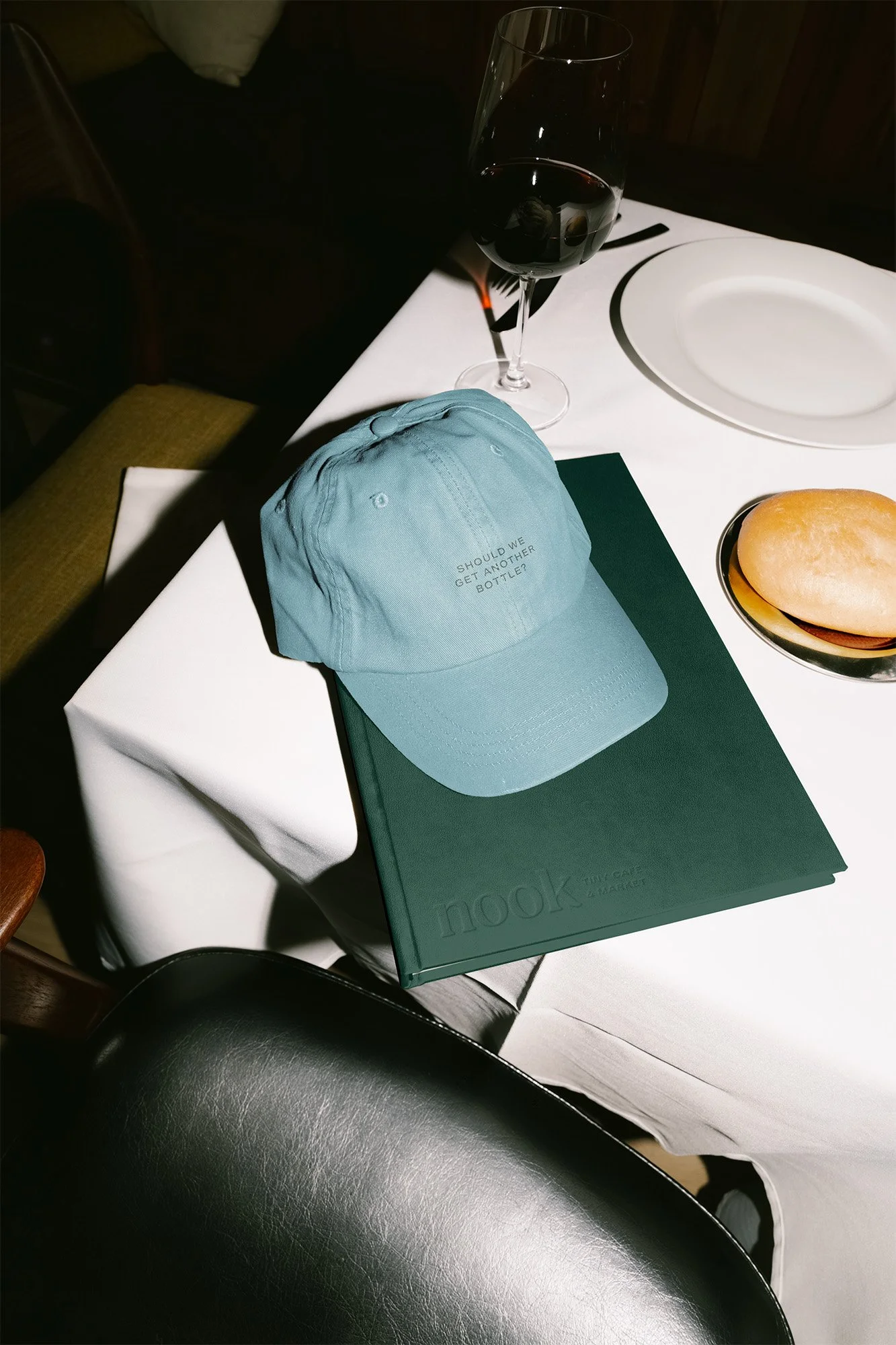 A dining table set with a glass of red wine, a book titled 'nook,' a light blue cap with the text 'SHOULD WE GET ANOTHER BOTTLE?,' a bread roll on a small plate, and a white empty plate.