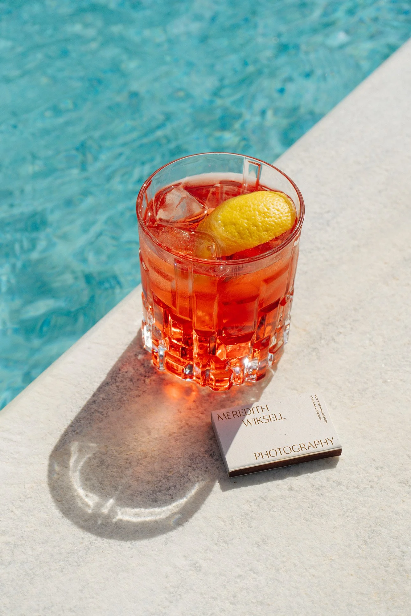 A glass of pink cocktail with ice and a lemon wedge, placed on a white surface near a pool, with a small white card reading 'Meredith Wiksell Photography' next to it.