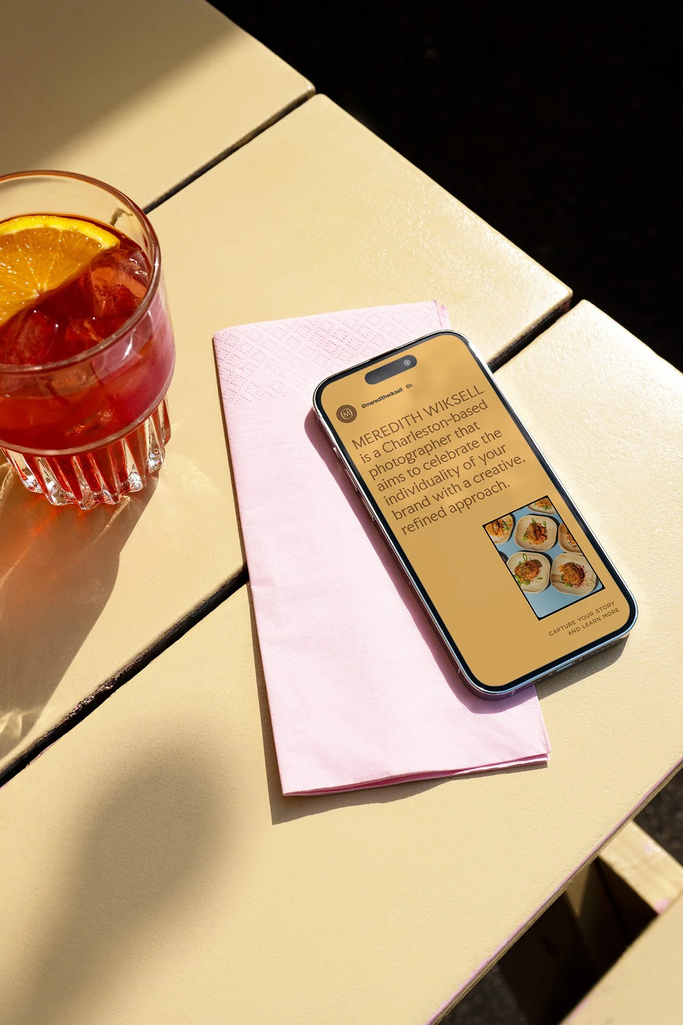 A smartphone with a yellow background on the screen, displaying a profile of Meredith Wicksell, a Charleston-based photographer, on a pink napkin, next to a glass of iced tea with lemon slices on a beige table.