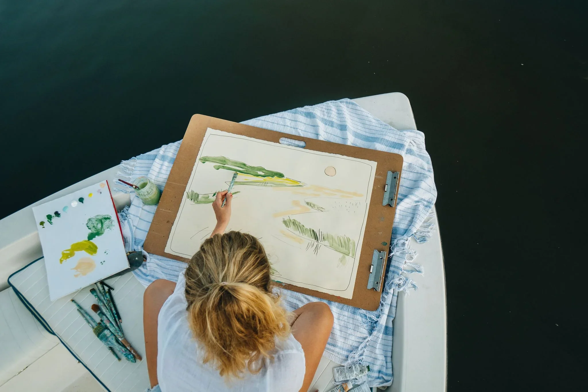 A woman sitting on a boat, painting a landscape on a large sheet of paper attached to a drawing board. The scene includes water, trees, and a setting sun. The boat's surface holds painting supplies like brushes, a palette, and a jar.