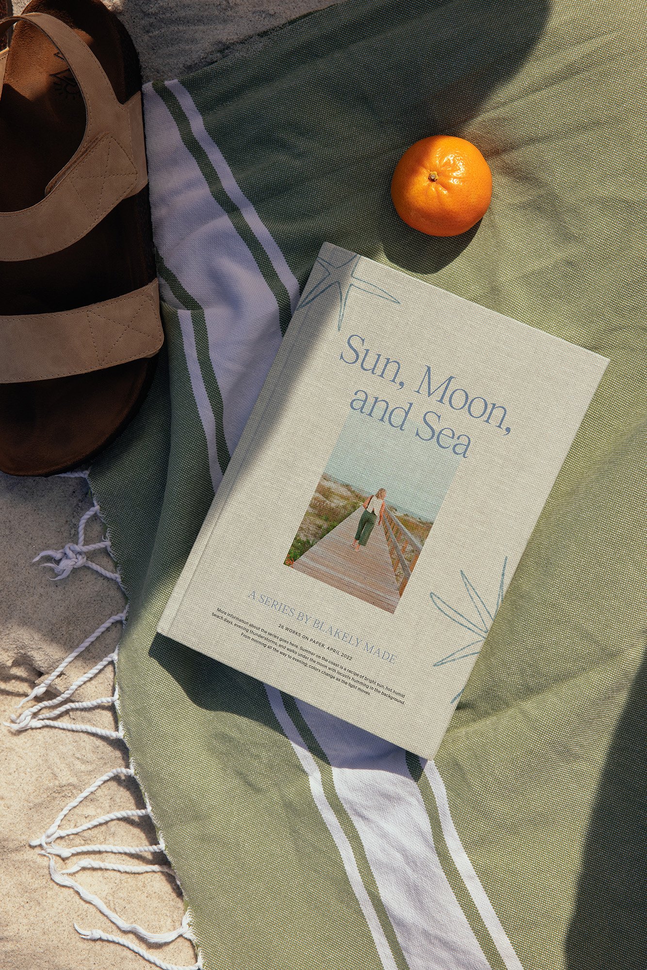 A beach blanket with a book titled 'Sun, Moon, and Sea,' an orange, a pair of sandals, and part of a bag, set on sandy ground with sunlight casting shadows.