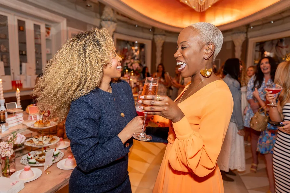 Two women smiling and chatting at a celebration, holding glasses of sparkling wine, with a decorated table of desserts in the background.