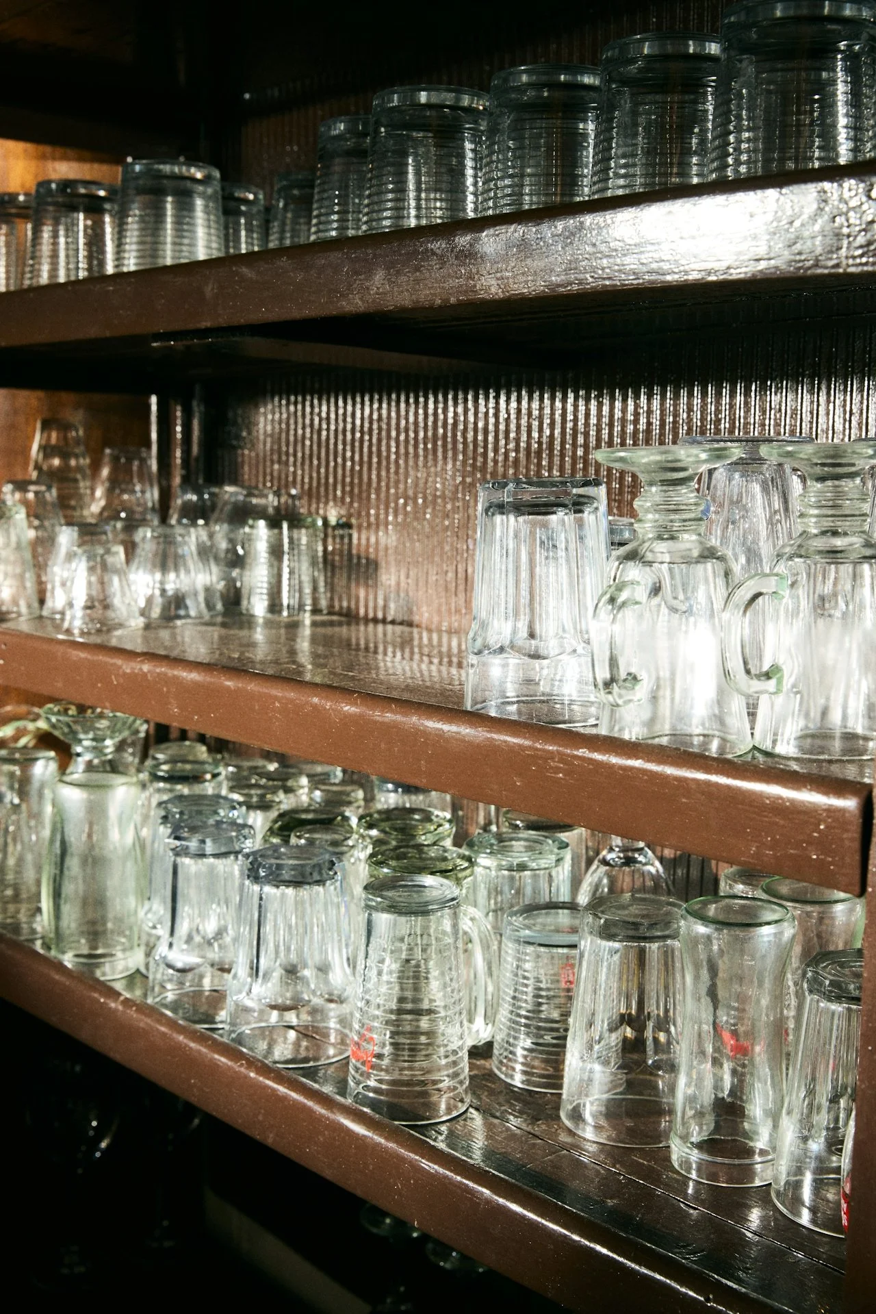 Multiple glass cups and mugs stacked on wooden shelves in a restaurant or cafe setting.