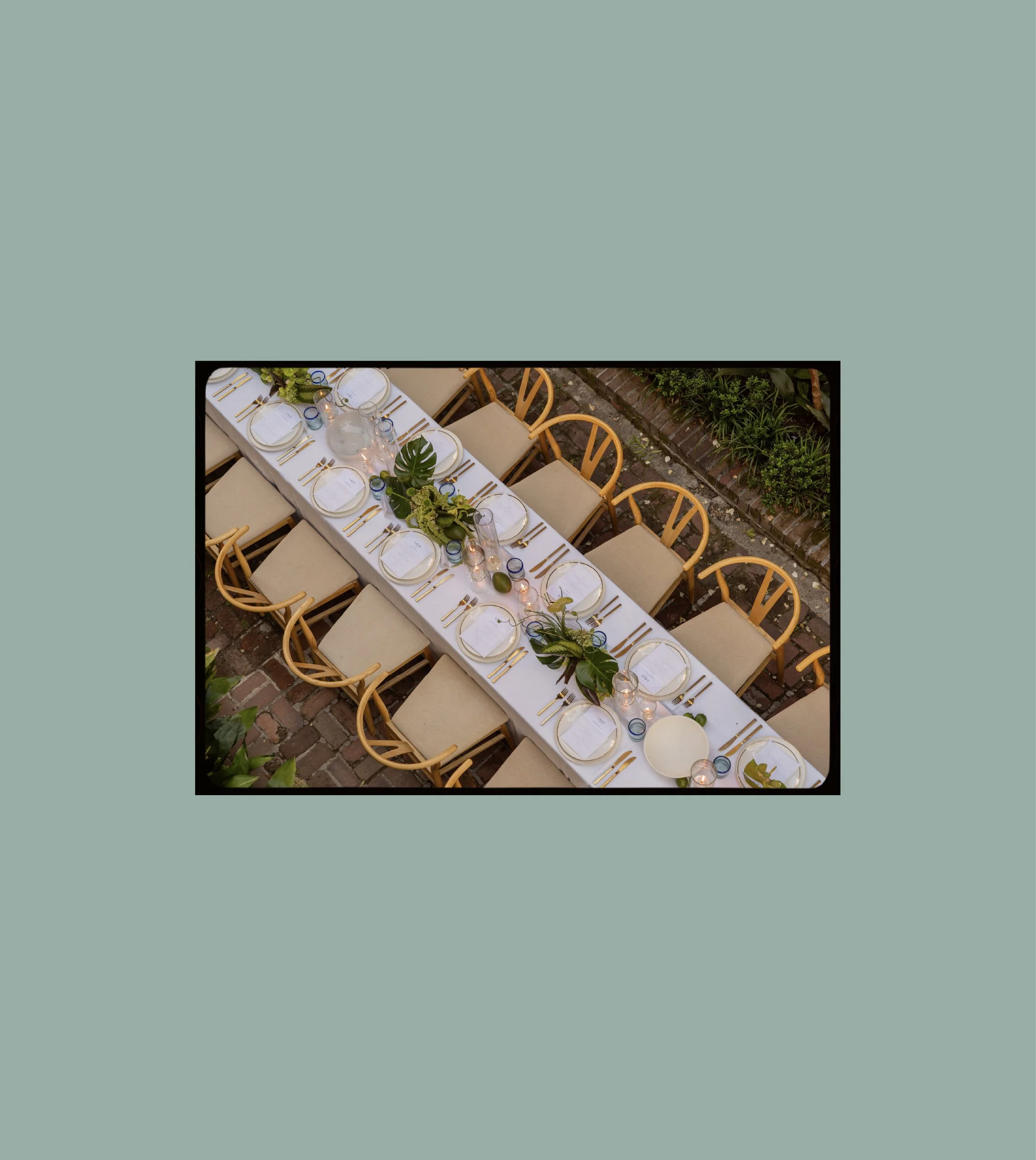 Outdoor dining table set for a formal meal with white plates, gold utensils, glassware, candles, and green foliage centerpieces.