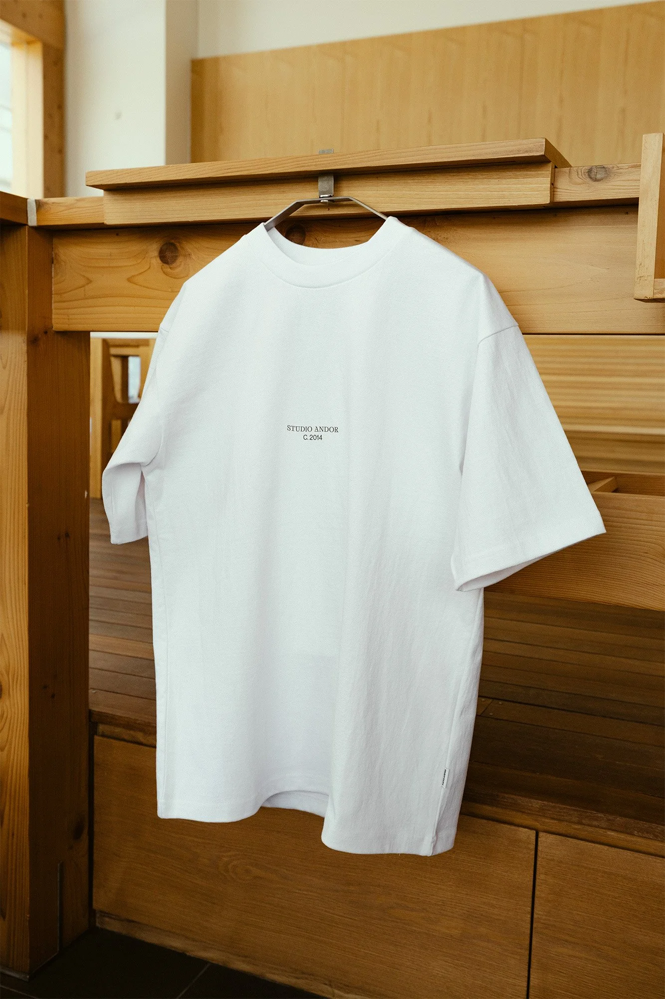 White T-shirt hanging on a hanger against a wooden background with small text in the center that reads 'STUDIO ANDOR C.2014'