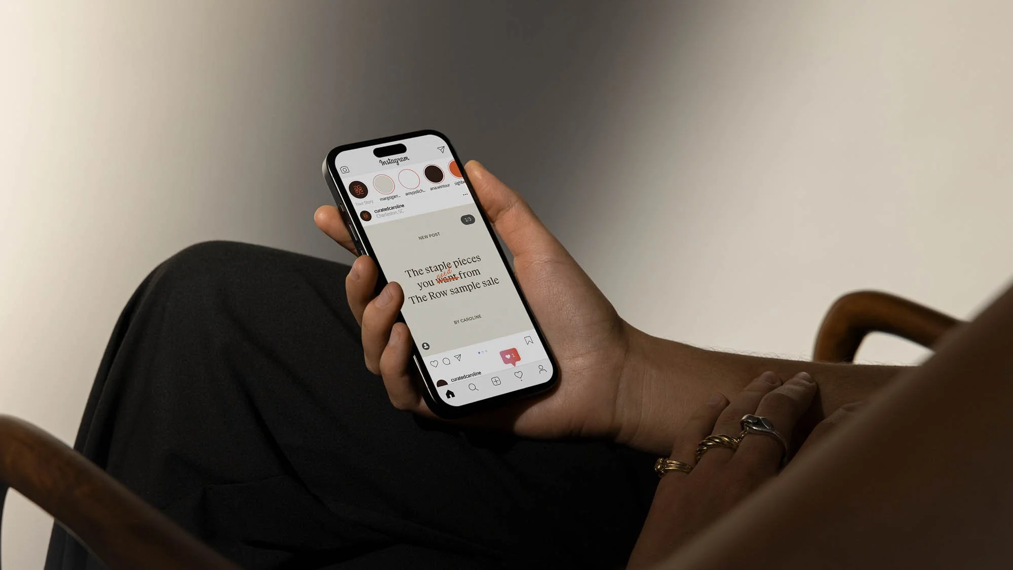 Person sitting on a wooden chair holding a smartphone displaying an Instagram post about a sample sale, with their other hand resting on their lap. The person is wearing dark clothing and gold rings.