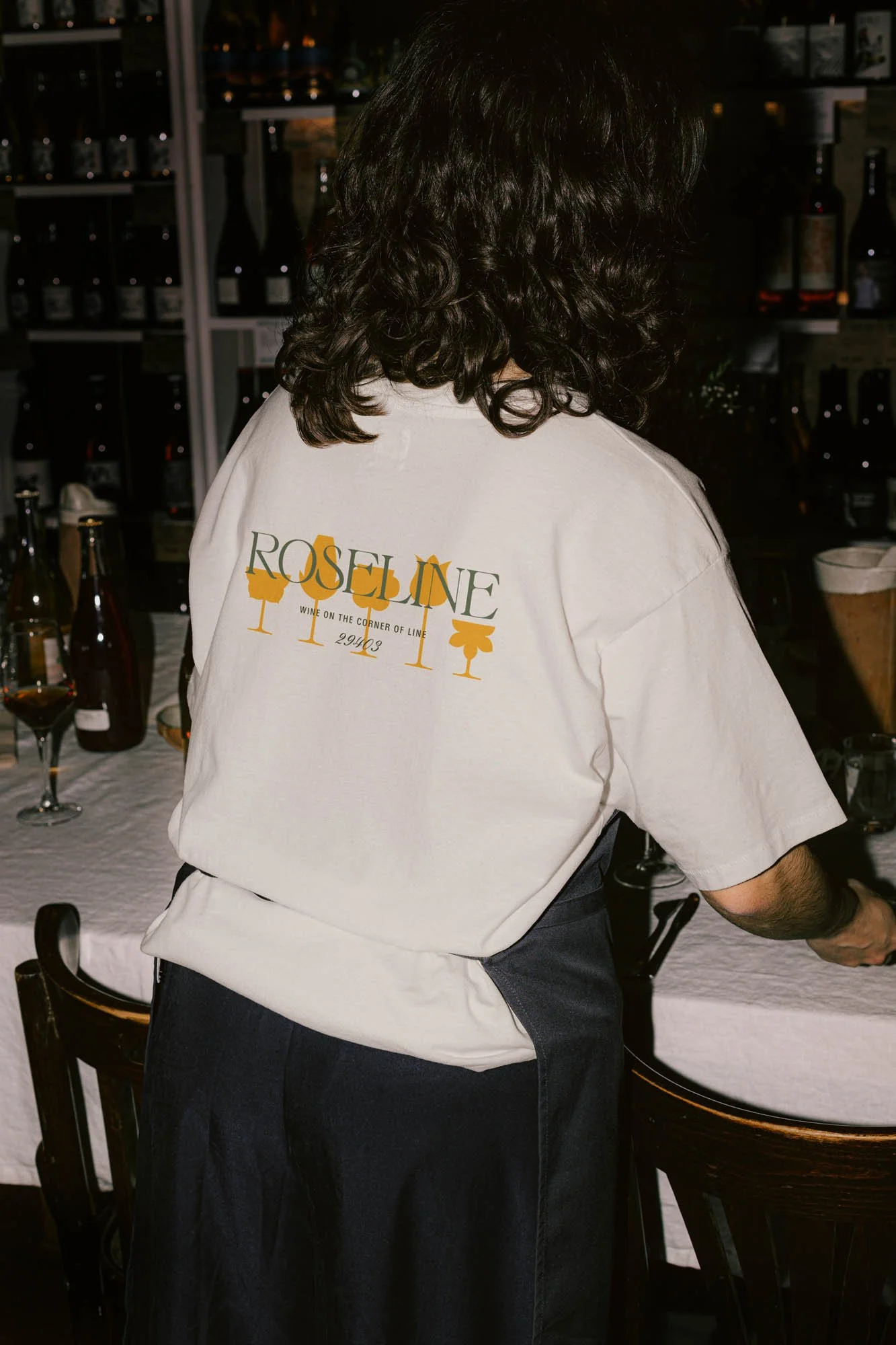 Person with long dark hair wearing a white "Roseline" branded t-shirt and black apron, standing near a set table with wine bottles and glasses, in a dimly lit setting.