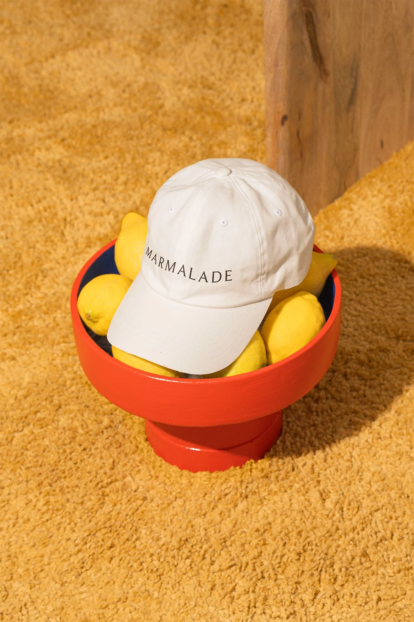 A red bowl filled with yellow lemons, with a white cap labeled 'MARMALADE' resting on top, placed on a textured beige carpet next to a wooden surface.