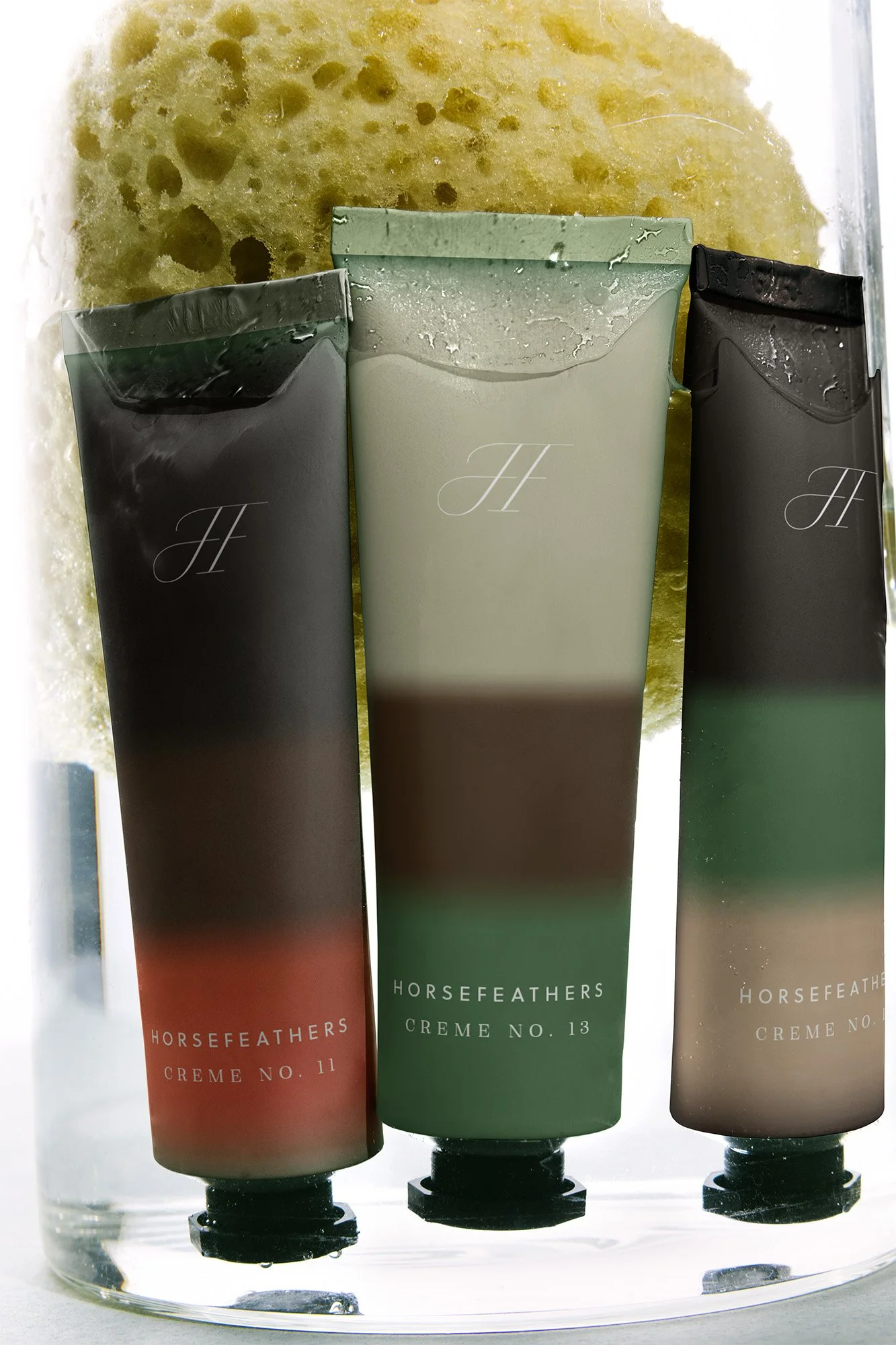 Three tubes of HorseFEATHERS Creme No. 11, No. 13, and an unidentified shade, standing upright behind a yellow sponge in a clear container.