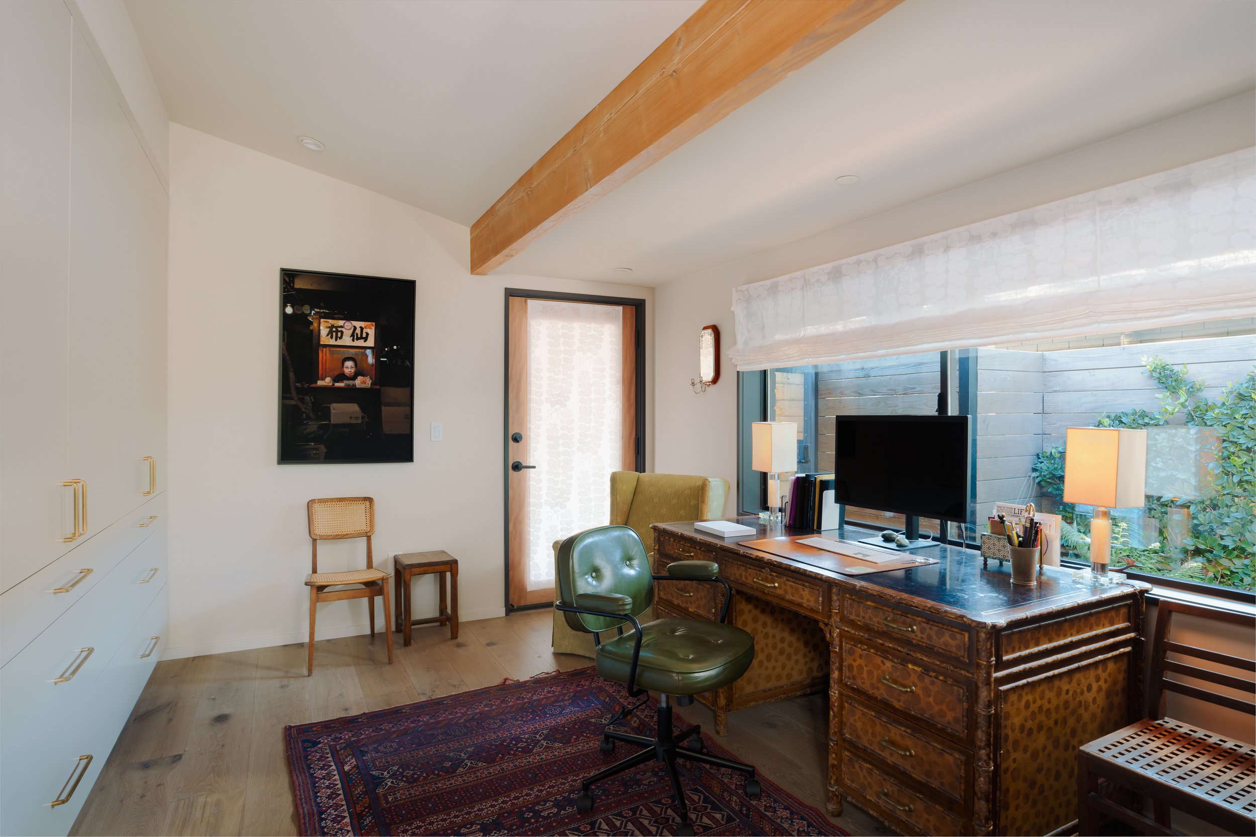 Home office with vintage wooden desk, green swivel chair, computer monitor, table lamps, and window with white blinds, beige walls, hardwood floor, and decorative rug.