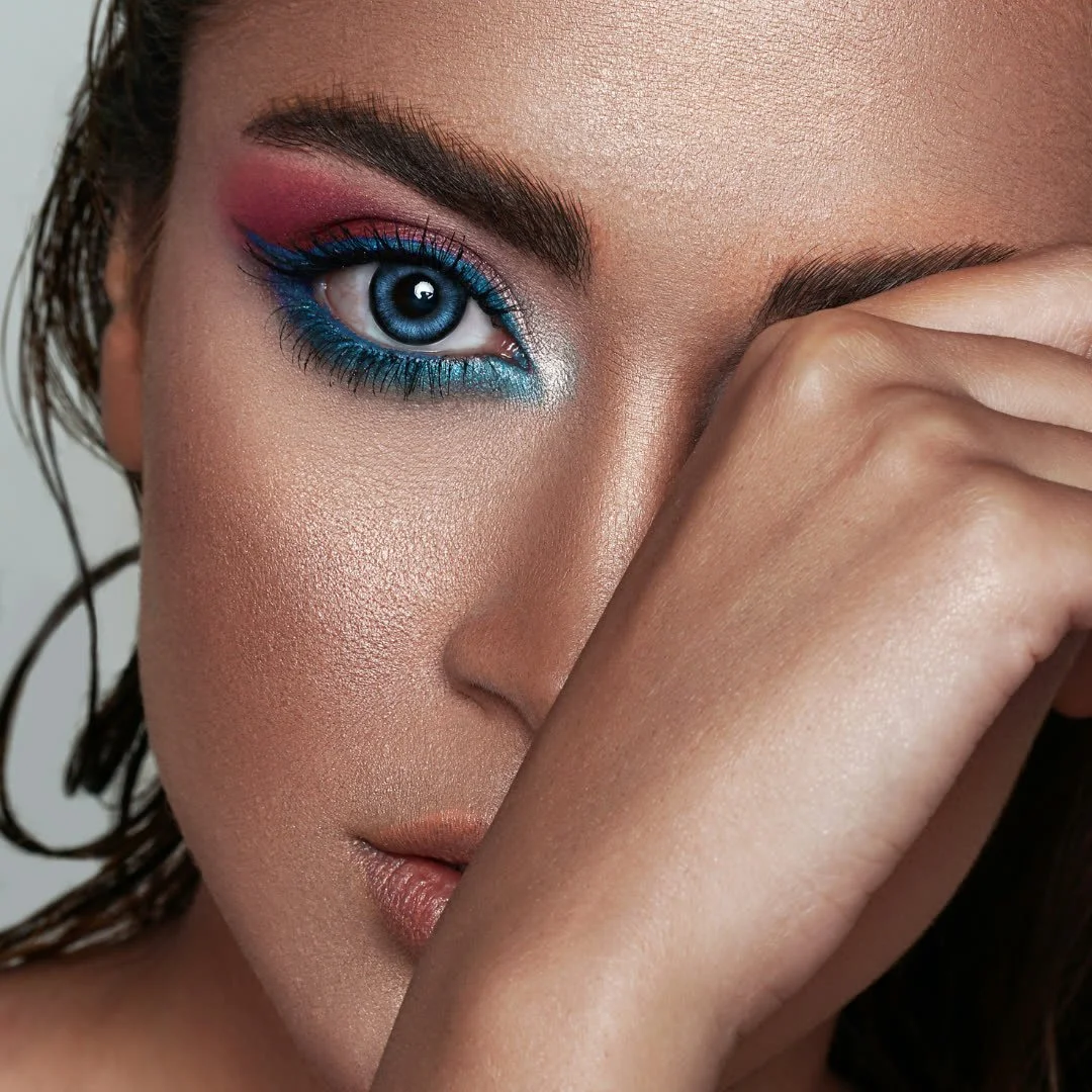 Close-up of a woman with vibrant makeup, featuring bold blue and pink eyeshadow, long lashes, and her eye partially covered by her hand.
