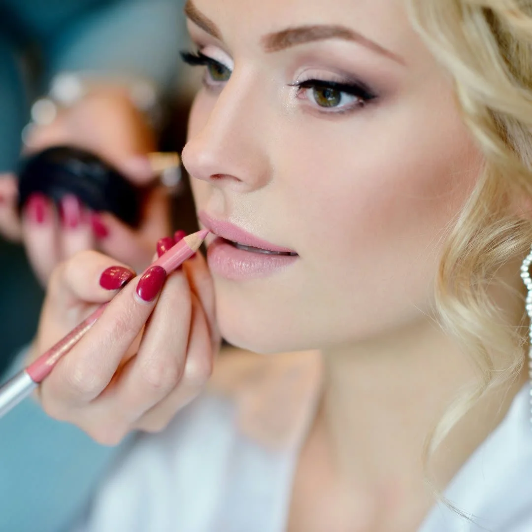 Close-up of a woman with blonde hair getting her makeup applied, focusing on her face as a makeup artist uses a pink lip liner near her lips.