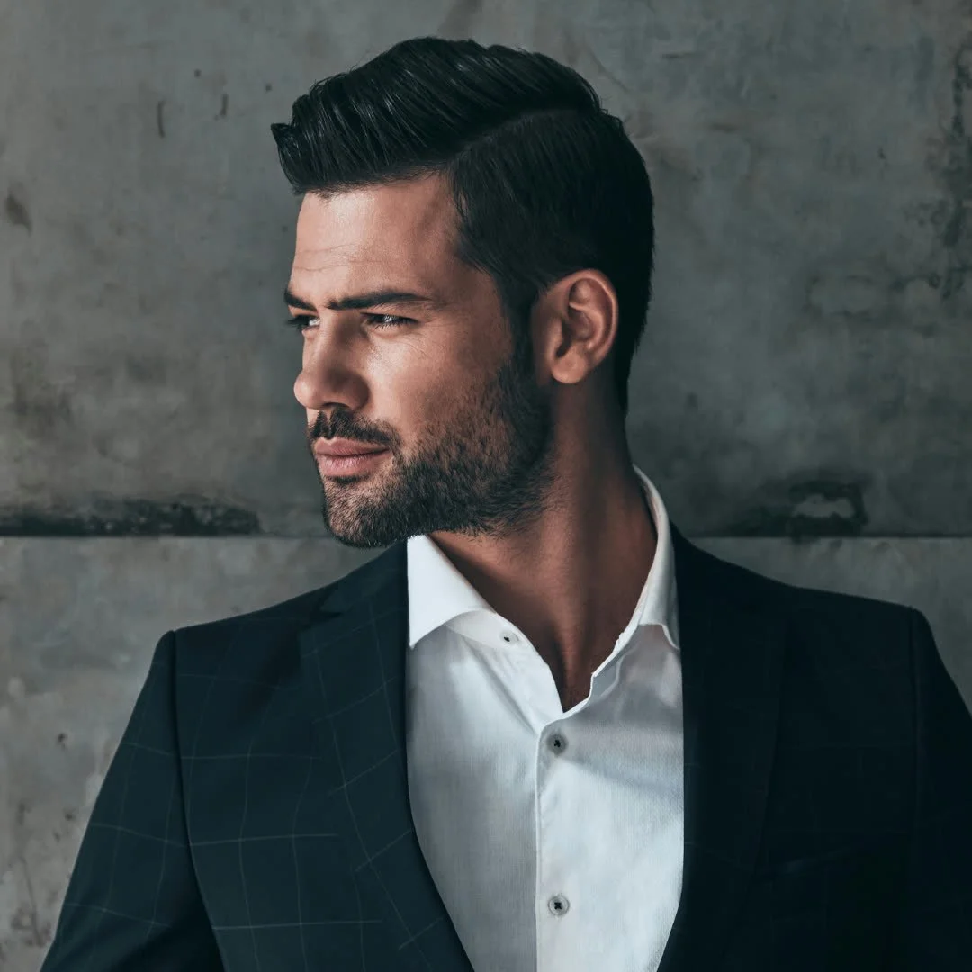 A man with dark hair and a beard looking to the side, dressed in a black suit jacket and white shirt, standing against a gray concrete wall.
