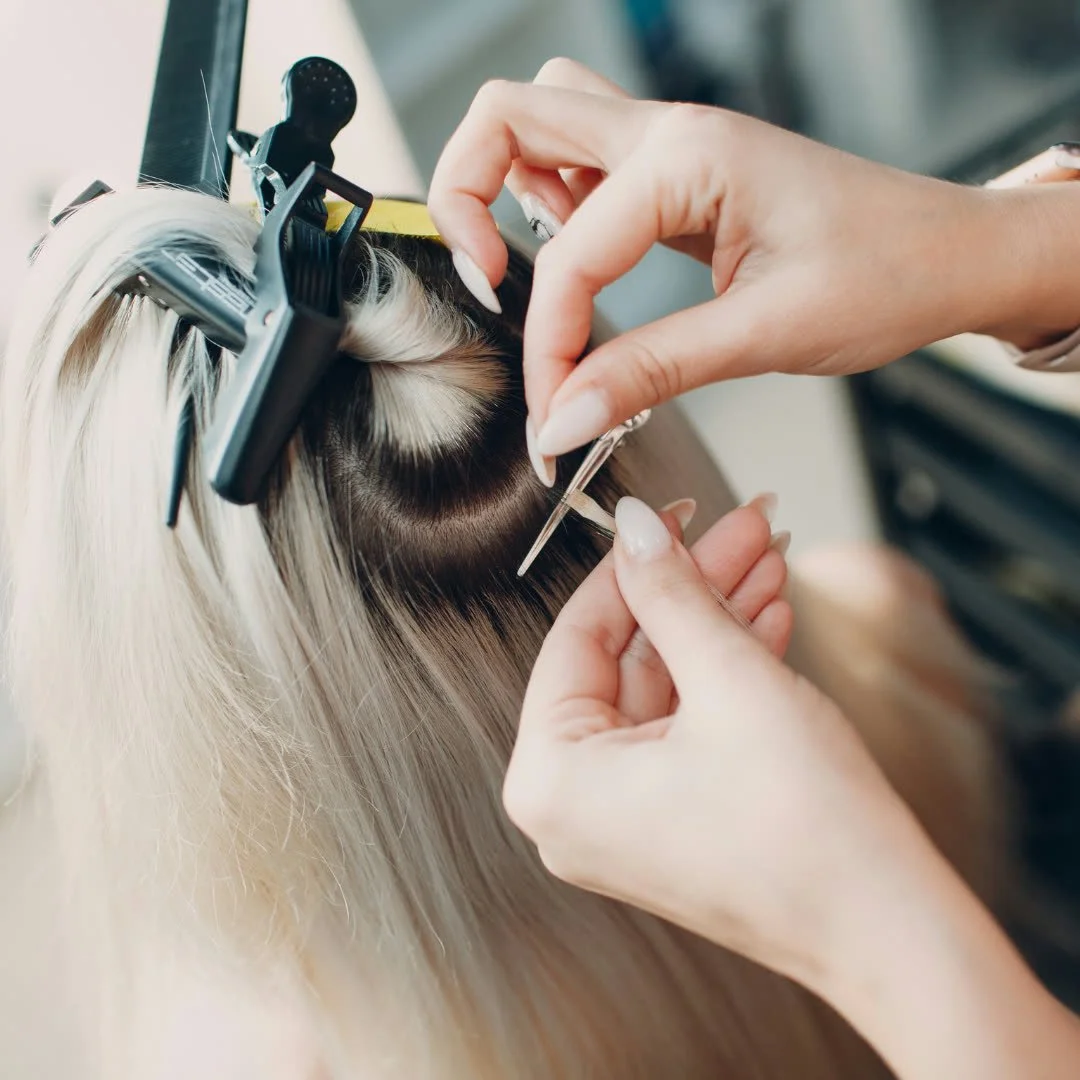 Close-up of a hairstylist applying hair extensions or doing a hair weave on a client with blonde and brunette hair, using a needle and thread.