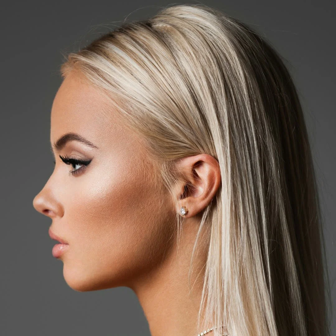 Profile of a woman with long, straight blonde hair, wearing diamond earrings and subtle makeup against a gray background.