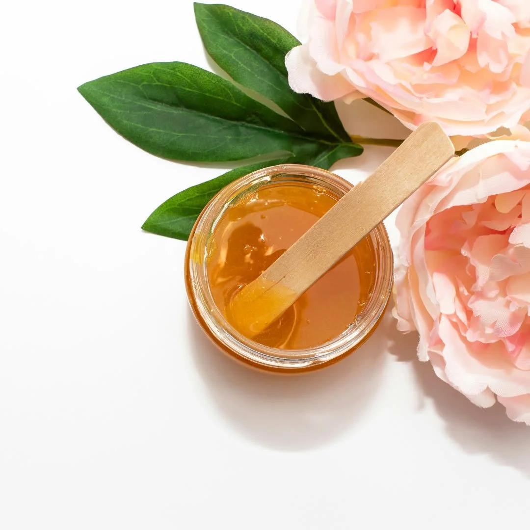 Jar of honey with a wooden honey dipper, pink peony flowers, and green leaves on a white background.