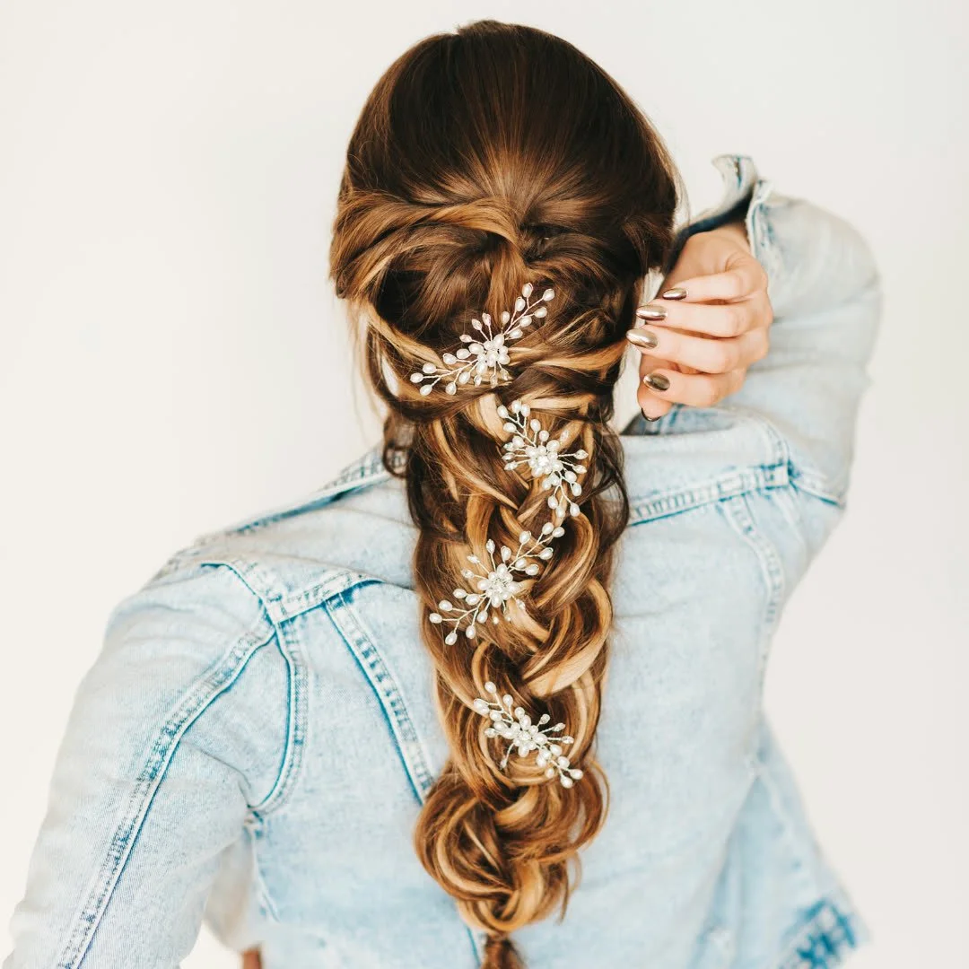 Close-up of a woman with long braided hair decorated with white floral hairpiece, wearing a light blue denim jacket, standing against a plain white background.