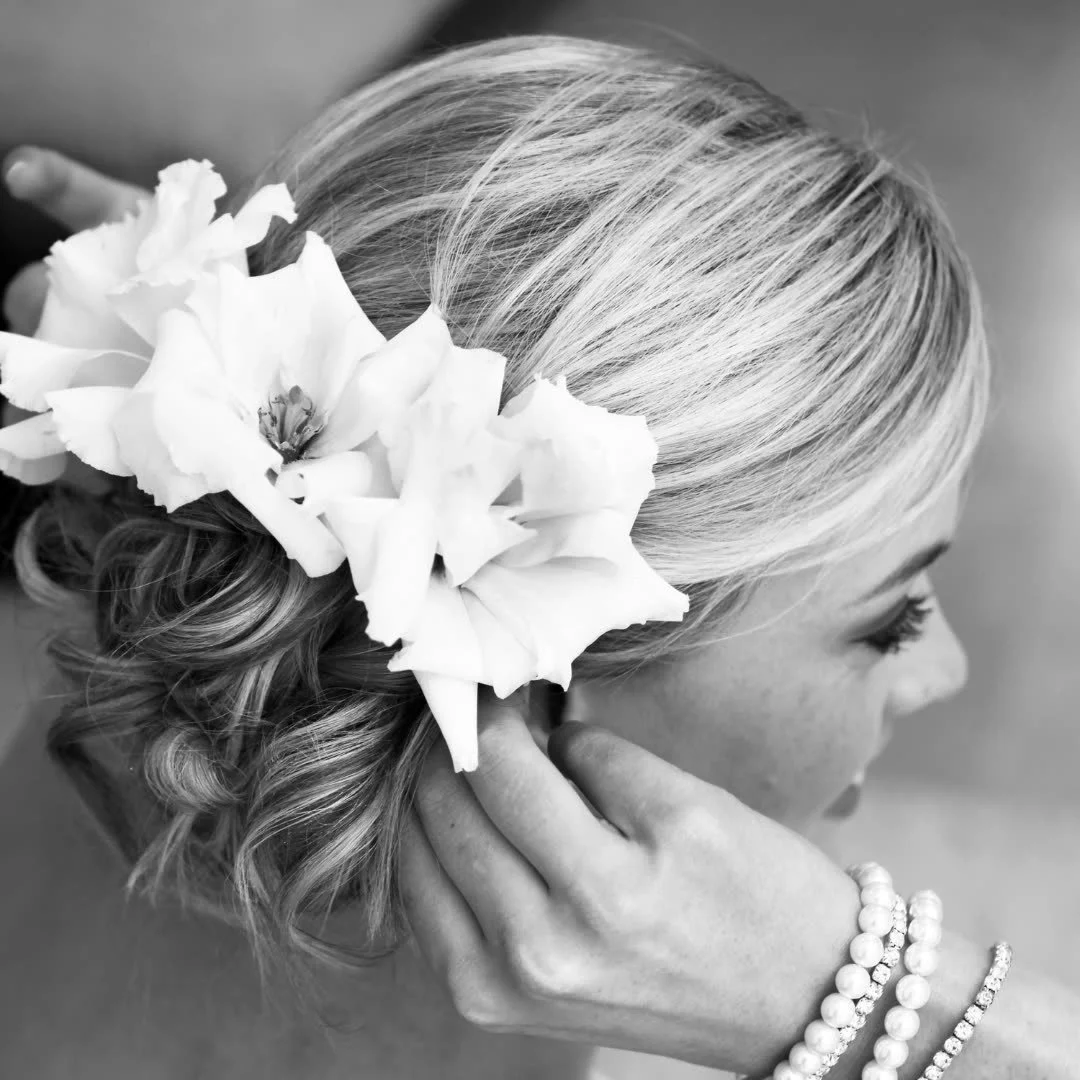 A woman with blonde hair decorated with large white flowers, touching her earring, wearing pearl bracelets, in a black and white photo.