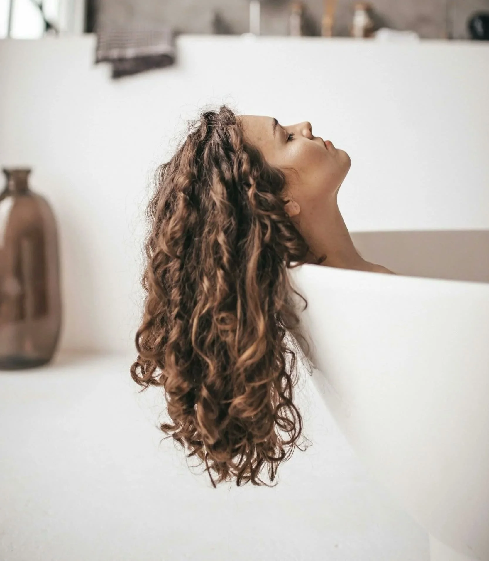 A woman with long, curly hair reclining in a bathtub with her eyes closed, in a modern bathroom setting.