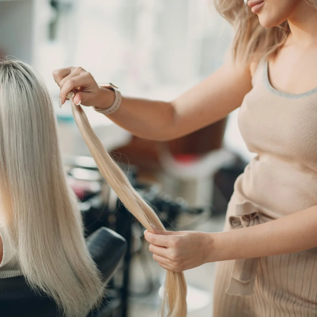 A woman with blonde hair styling a client's long blonde hair in a salon.