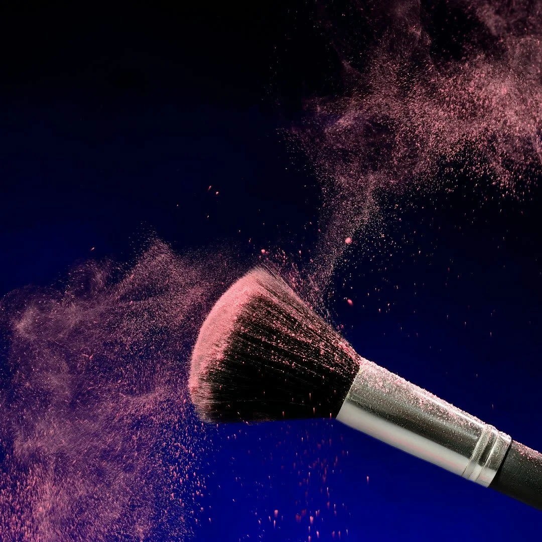 A makeup brush disperses pink powder into the air against a dark background.