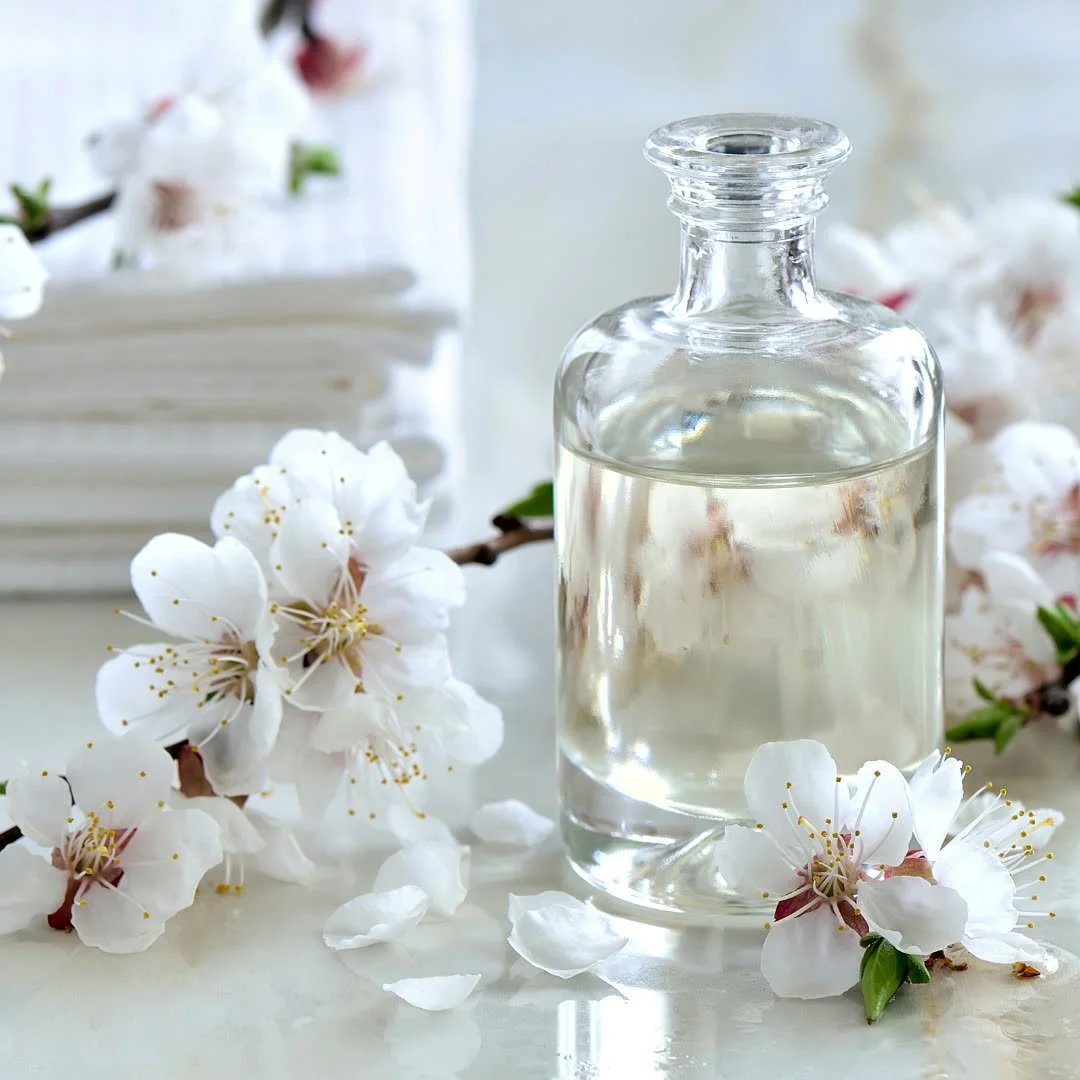 A glass bottle of clear liquid surrounded by white cherry blossoms and some petals on a white surface.
