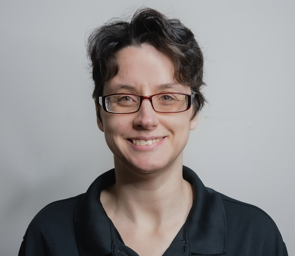 Balanced Chiropractic with short dark hair and glasses smiling, wearing a black collared shirt, against a neutral background.