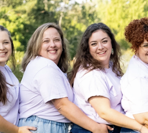 The Village Doulas Columbus, OH standing in a line outdoors, each wearing a light purple shirt and smiling. The background features greenery.