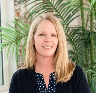 Columbus, OH Birth Doula & Childbirth Educator Caroline Pitts smiling with long blonde hair, wearing a navy blue polka dot blouse and cardigan, standing in front of green palm plants indoors.