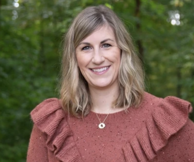 Leah Christie Columbus, OH Lactation Specialist IBCLC smiling outdoors in a rust-colored textured sweater with a ruffled shoulder detail, surrounded by green foliage background.