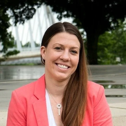 Melissa Foster Columbus, OH EMDR Mental Health Therapist wearing a coral blazer and white top is sitting outdoors, smiling. Trees and a bridge form the background. The setting appears to be a park.