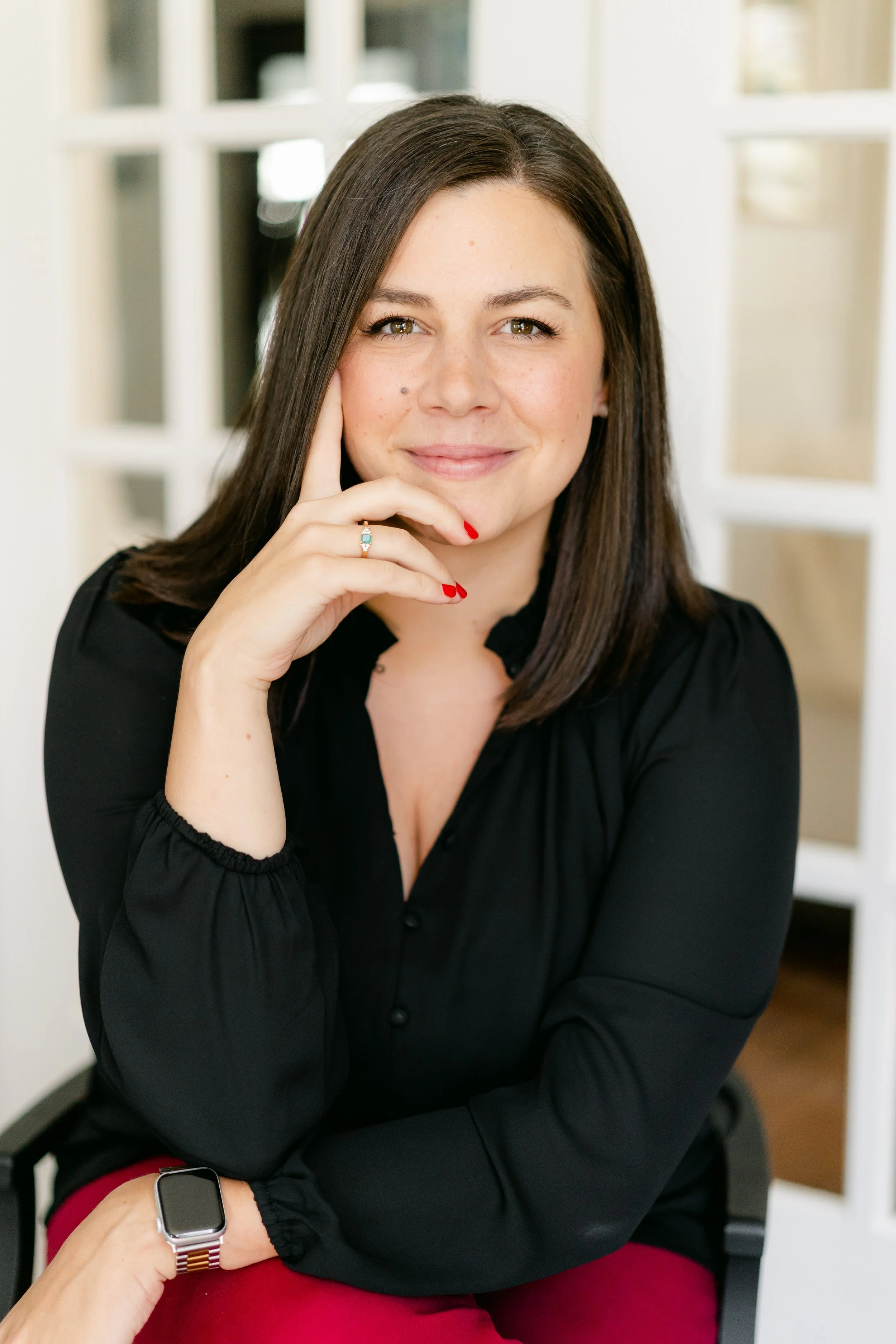 Sarah ODonnell Columbus, OH Sonder Mental Health Therapist with long dark hair wearing a black top and red pants, sitting in front of a background with windows, resting their chin on their hand.