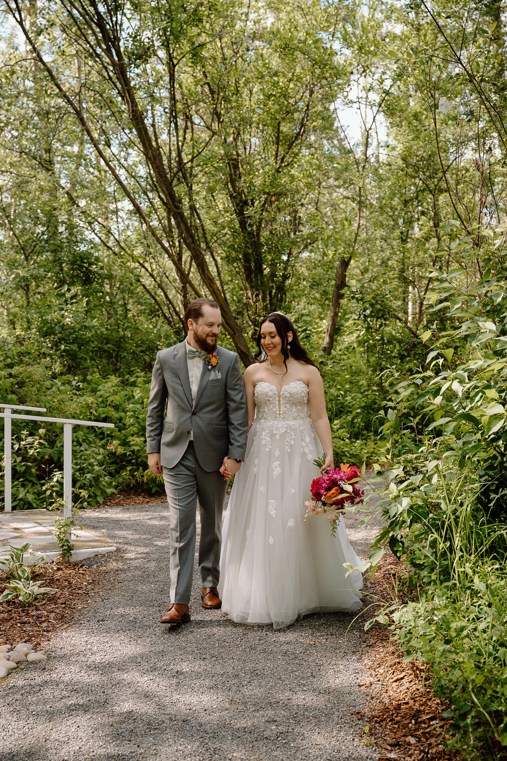 A bride and groom walking hand in hand outdoors at Sparrow Lane. The bride is holding a colorful bouquet and wearing a strapless white wedding gown, while the groom is in a gray suit with a bow tie and boutonniere.