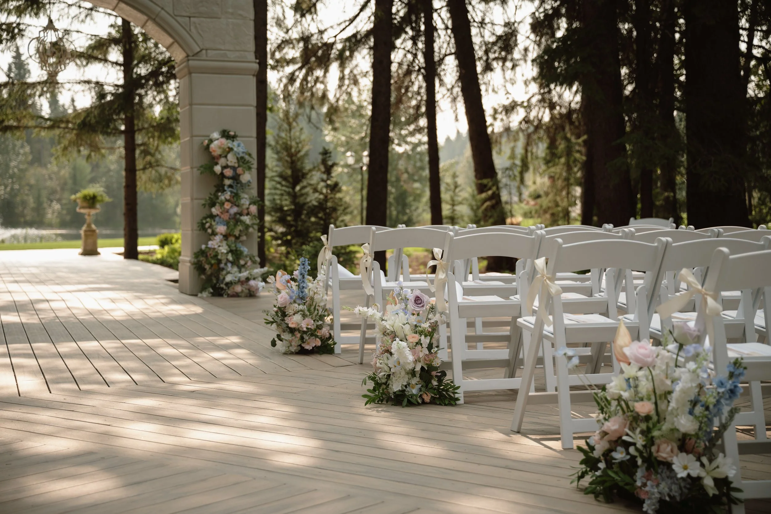 Wedding ceremony setup with white folding chairs decorated with cream ribbons, floral arrangements on the aisle, under a wooden arch overlooking a wooded landscape in Archway Manor, Red Deer, Alberta