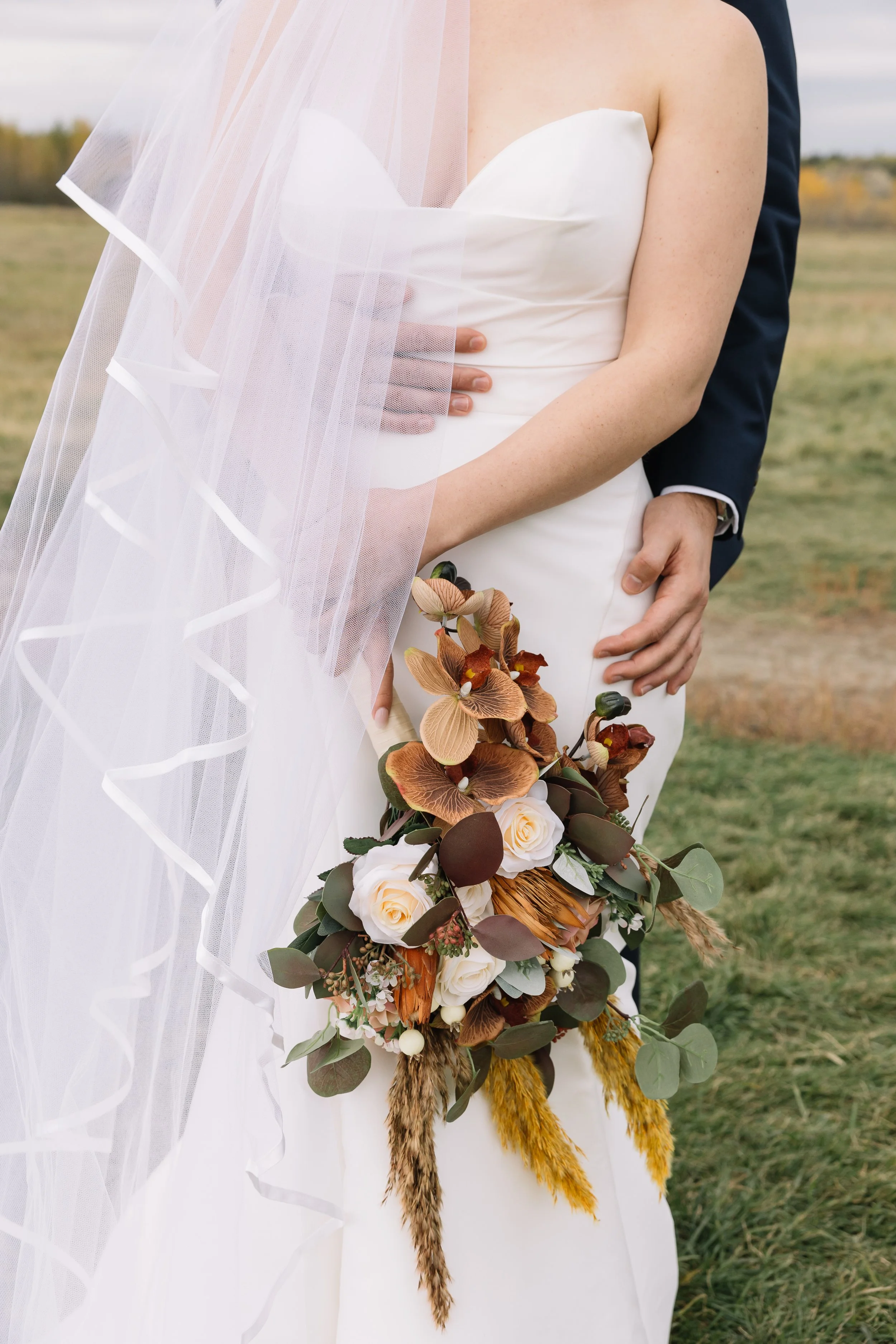 A bride in a white wedding dress and veil holding a bouquet of flowers, with a groom in a dark suit standing behind her, outdoors in a field.