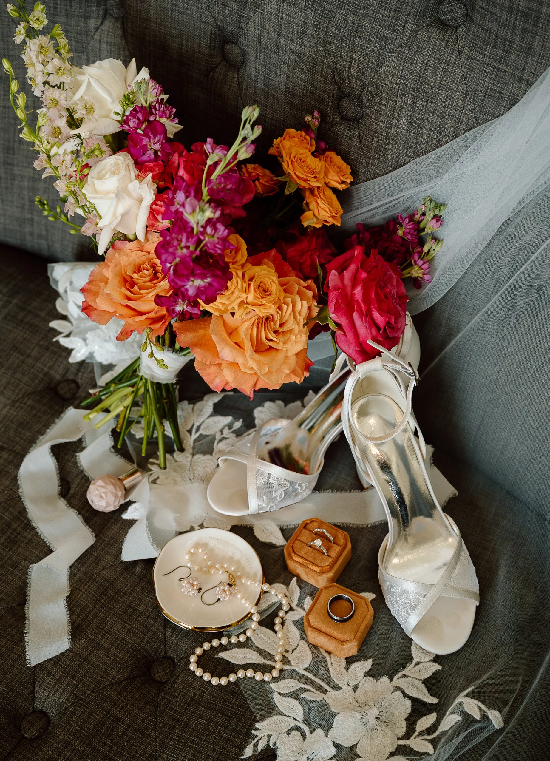 A wedding bouquet of colorful flowers, a pair of white lace high-heeled shoes, wedding rings in a box, pearl necklaces and earrings, placed on a floral tablecloth on a couch.
